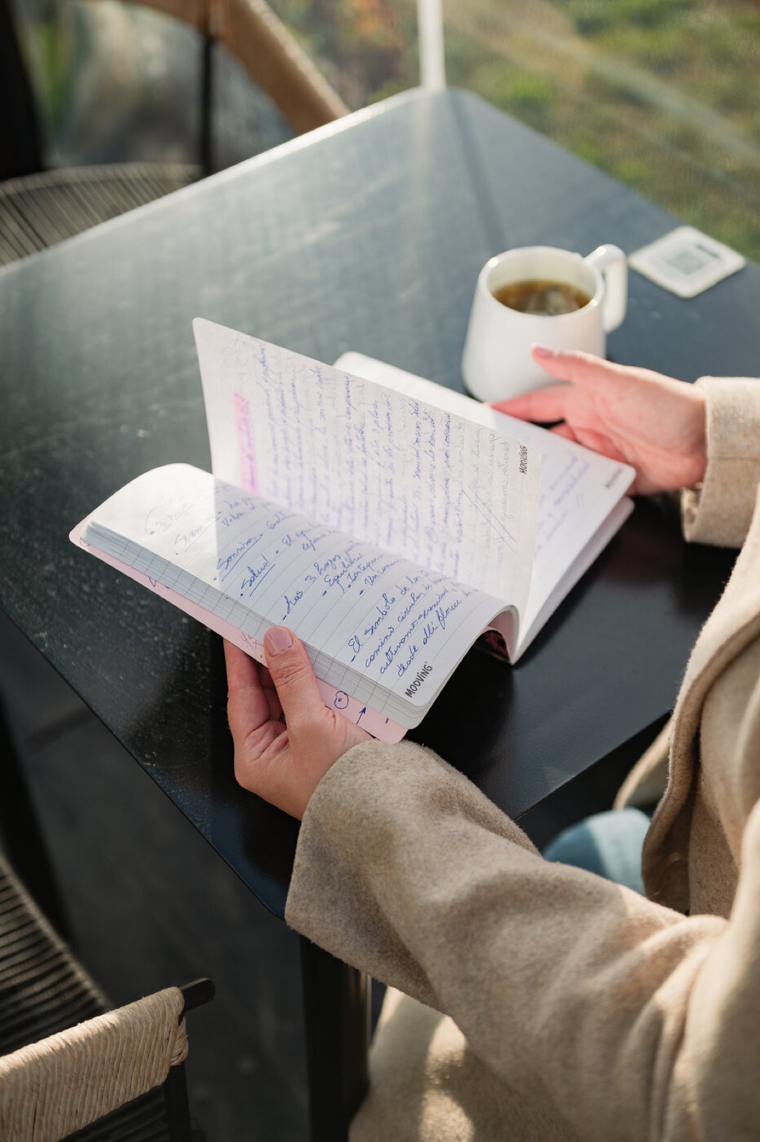 Manos sosteniendo un cuaderno escrito, con una taza de té cerca.