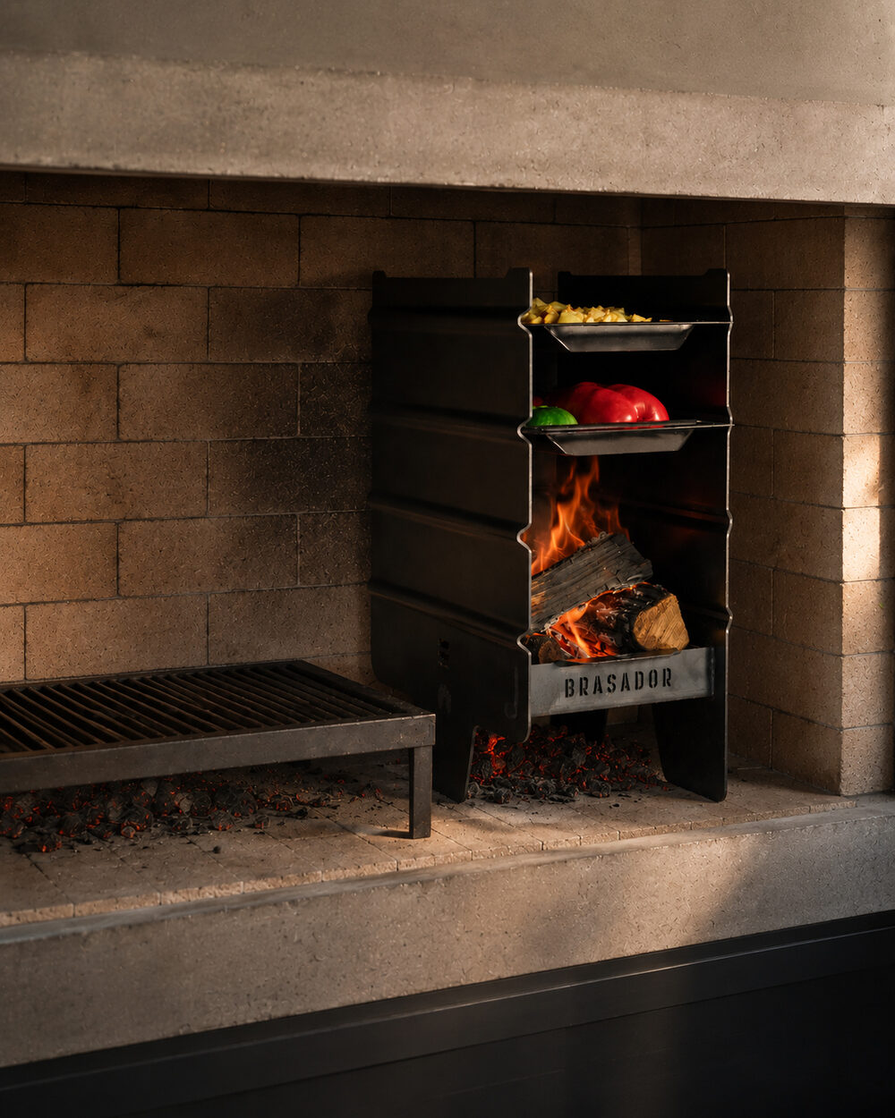 Brasador Esencial encendido, con las bandejas cargadas de verduras y el fuego adentro, junto a una parrilla de material
