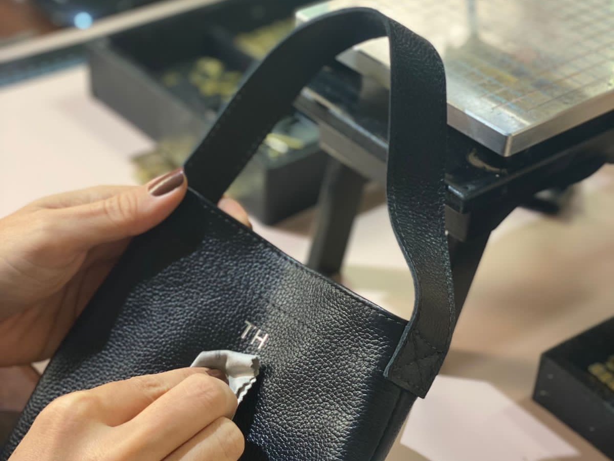 Mãos polindo o monograma TH recém-gravado em uma bolsa de couro preta, na bancada do ateliê, ao lado da prensa de hot stamping.