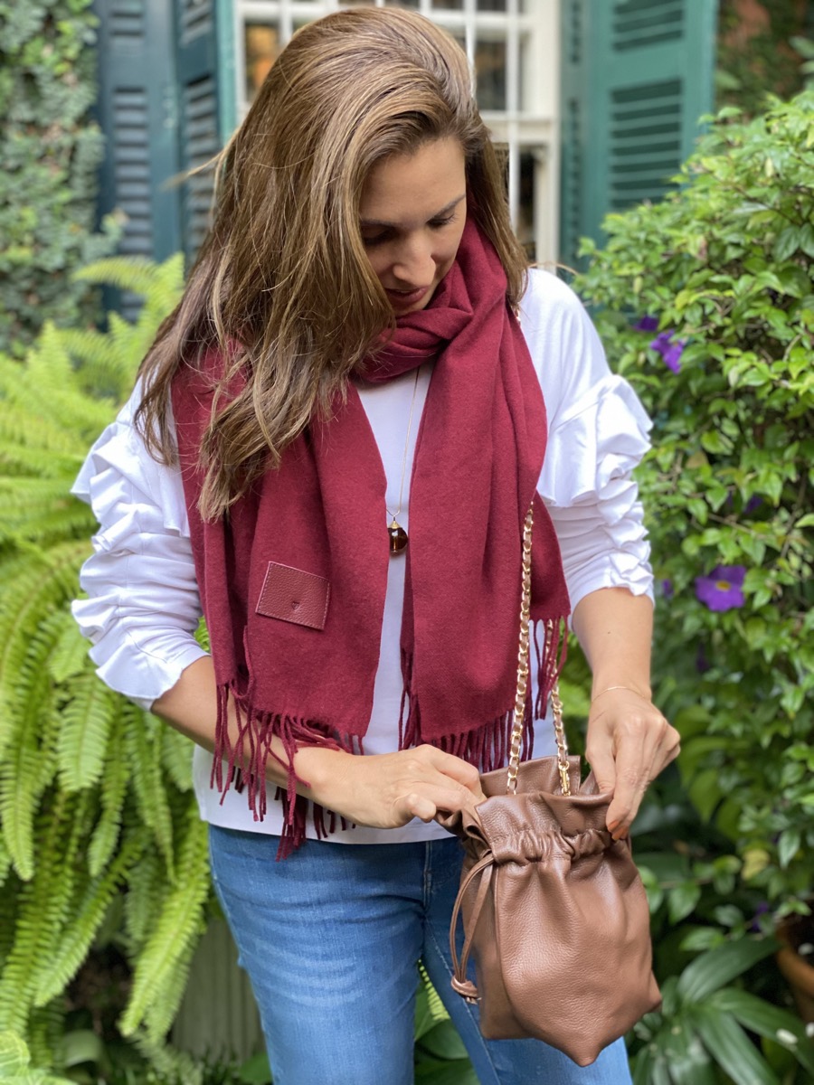 Lorena Simões, fundadora da Loeh, em um jardim, de blusa branca e echarpe vinho, abrindo uma bolsa saco de couro caramelo.