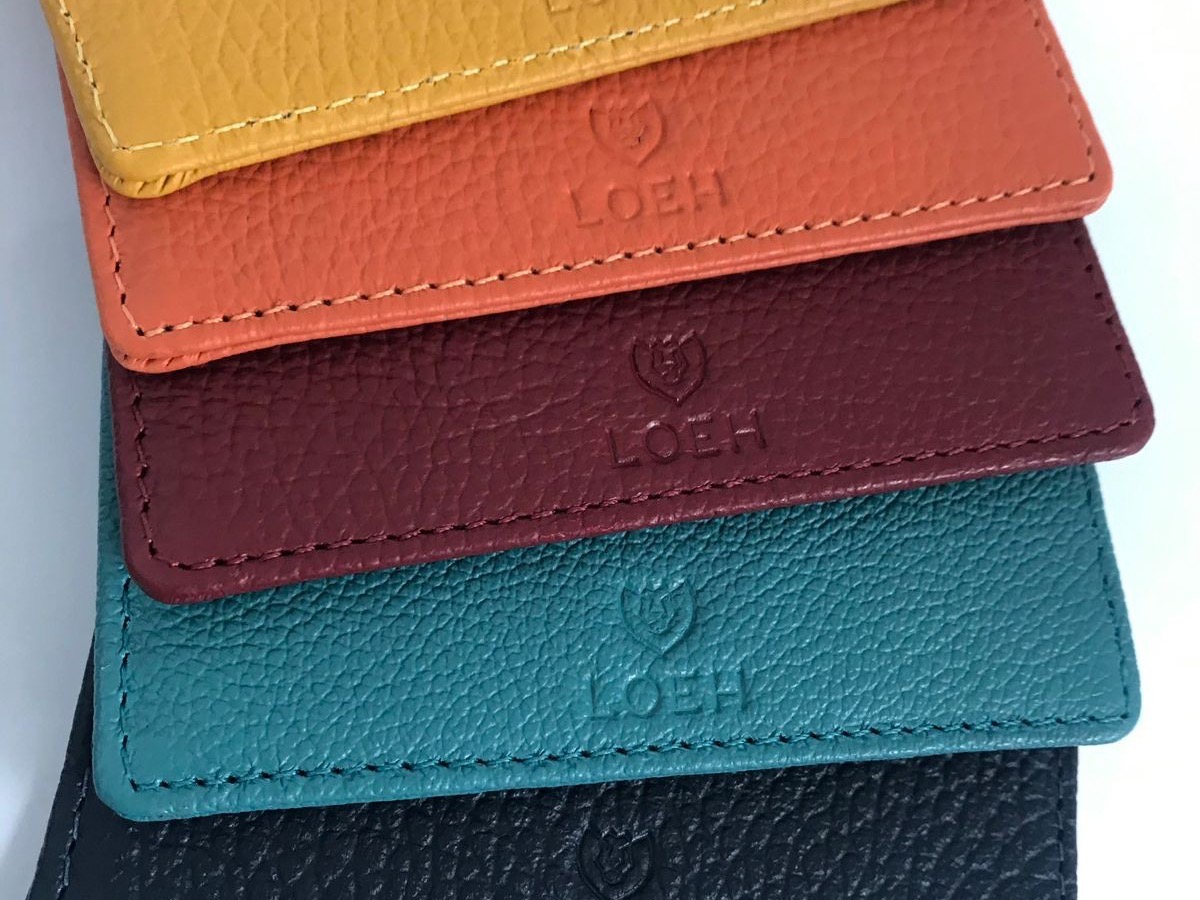 Cinco porta-cartões de couro empilhados em leque — mostarda, laranja, vinho, verde-petróleo e preto — cada um com o logotipo Loeh gravado.