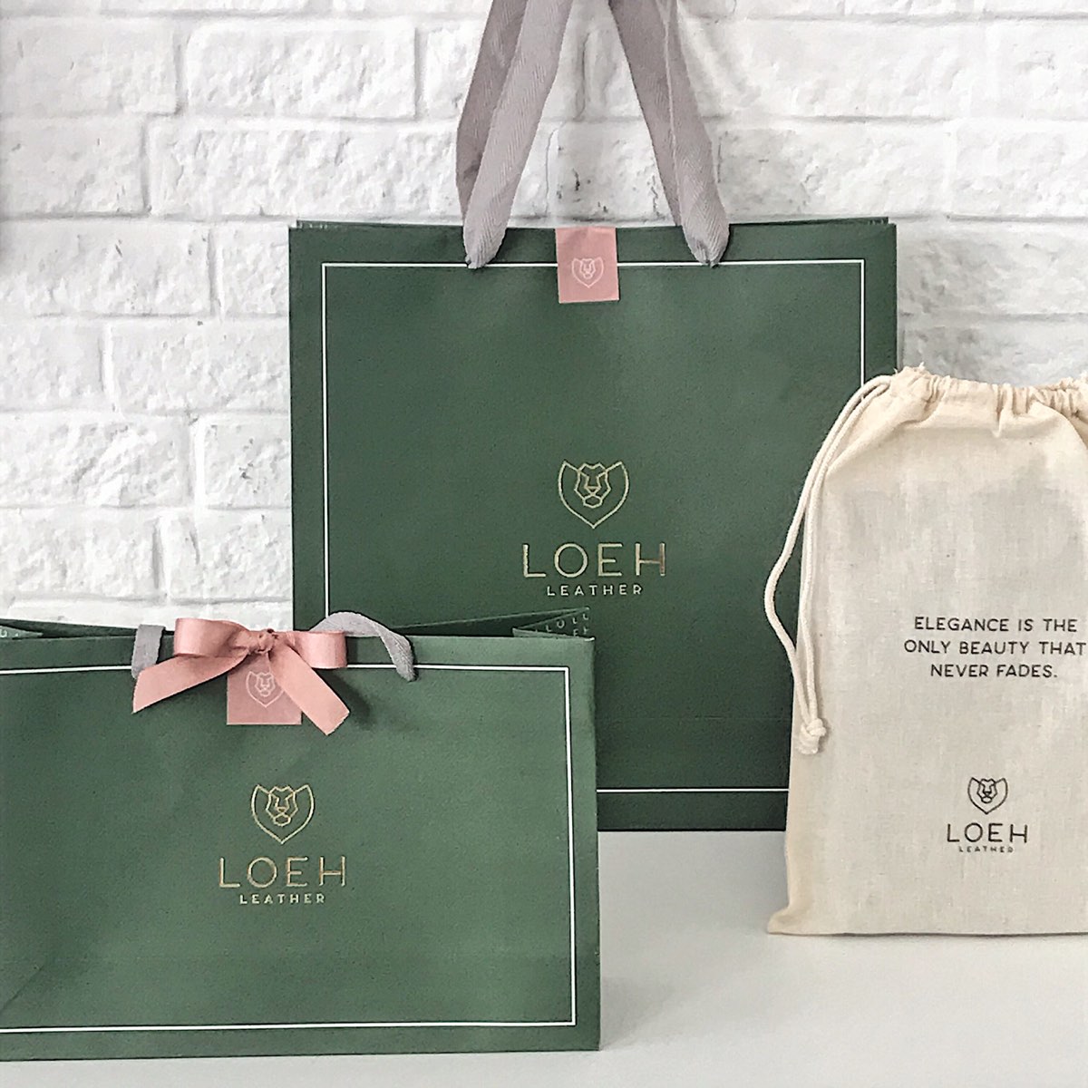 Caixas e sacolas verdes da Loeh com laço rosa, ao lado de um saquinho de algodão cru.