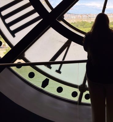 Silhueta de uma mulher de costas diante de um grande relógio de vidro com vista para a cidade.
